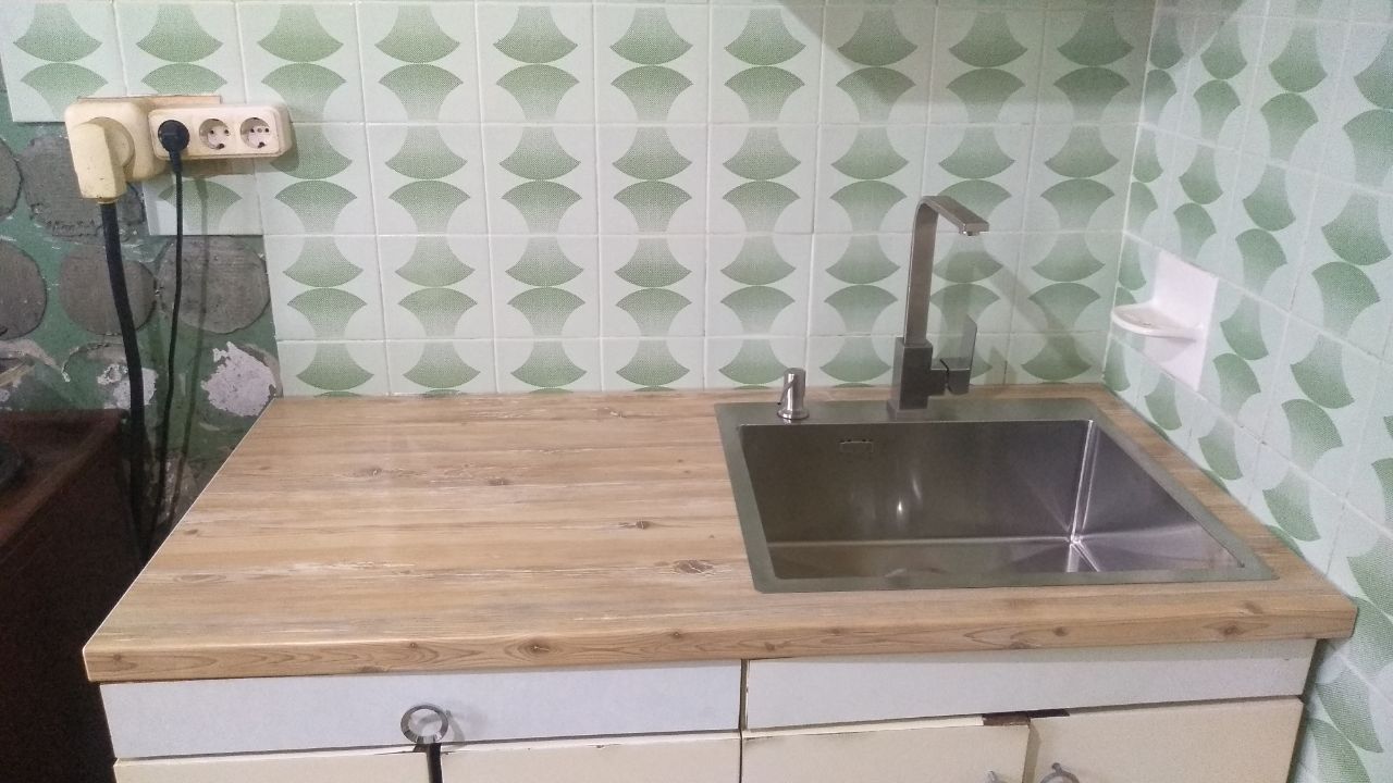 Small kitchen with a wooden counter, stainless steel sink, and green patterned wallpaper.