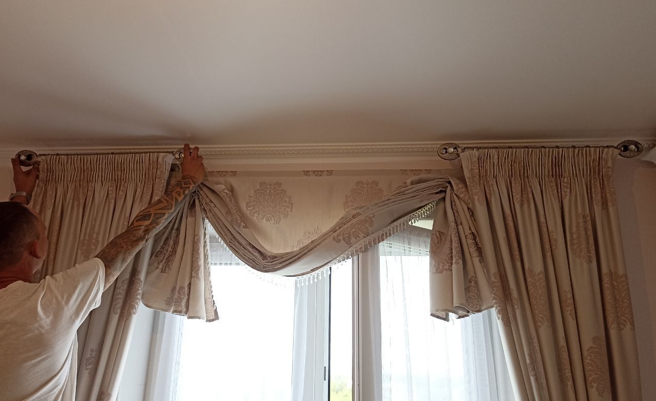 Person adjusting elegant cream curtains with a swag valance over a window.