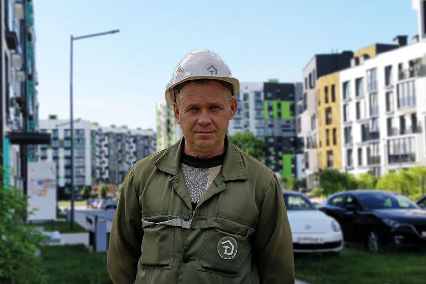 A construction worker in a hard hat and green jacket stands before modern apartment buildings.
