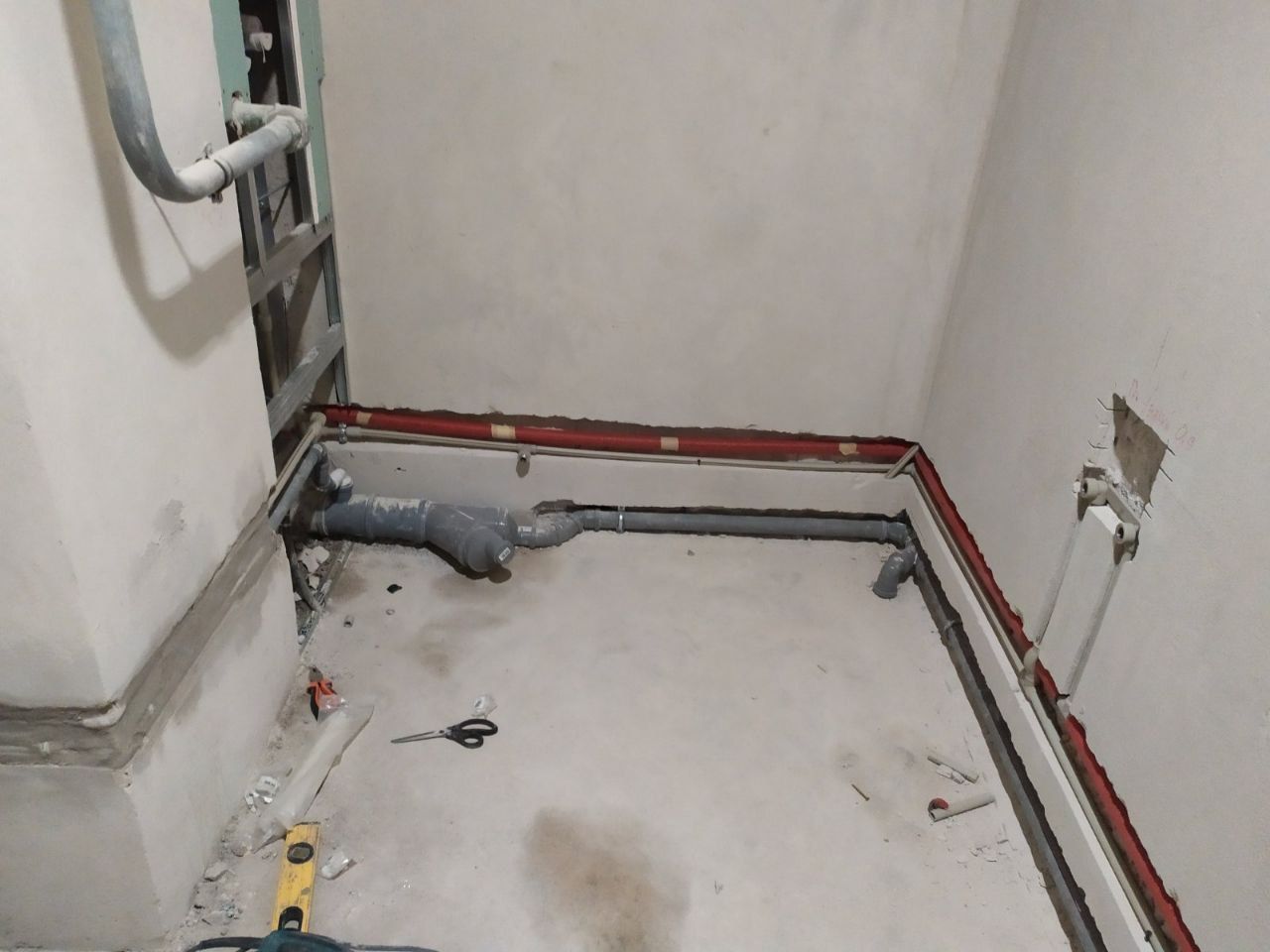 Unfinished bathroom with exposed grey plumbing pipes on concrete floor and bare white walls.