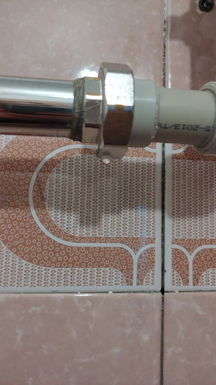 A water droplet drips from a leaking metal and plastic pipe connection against tiled wall.