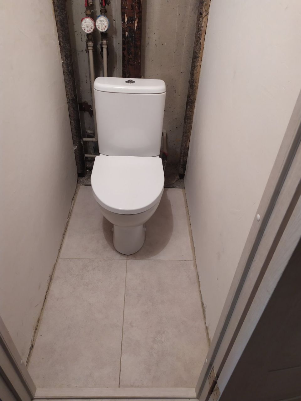 White toilet in a small, narrow bathroom with light tiled floor and visible pipes.