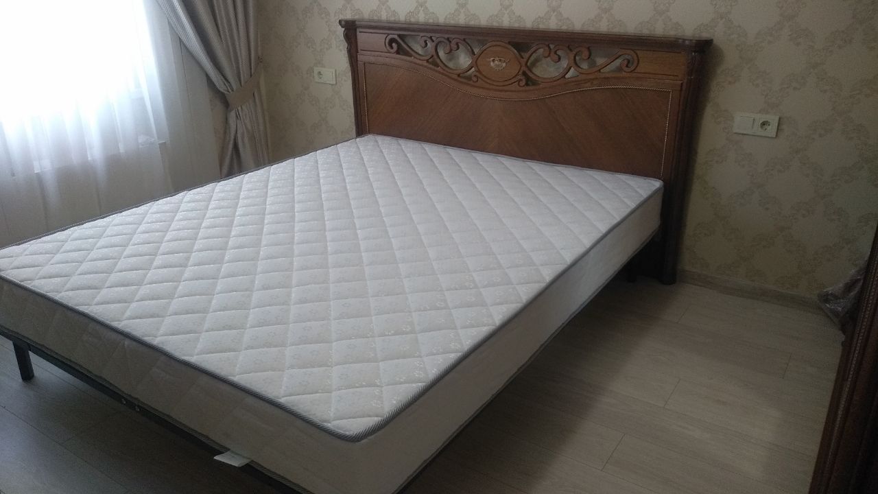 Bed with carved wooden headboard and white quilted mattress in a simple room.